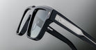 Close-up of stylish black eyeglasses with silver detailing on the temple arms, featuring a small engraved logo of a bird. The background is a soft gradient of grey.
