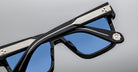 A close-up view of stylish black sunglasses with blue tinted lenses, featuring silver accents and a decorative emblem on the hinge, set against a neutral grey background.
