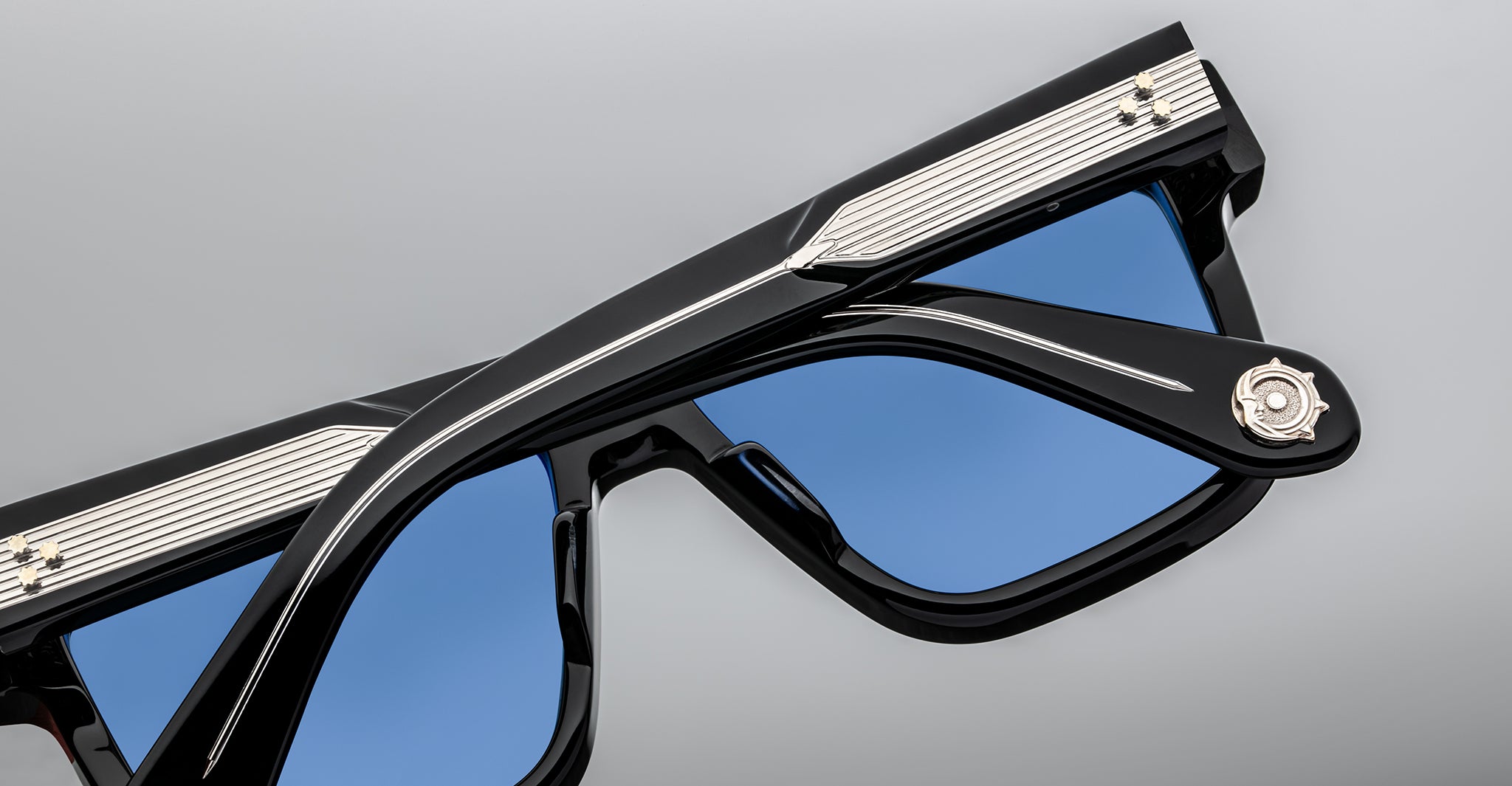A close-up view of stylish black sunglasses with blue tinted lenses, featuring silver accents and a decorative emblem on the hinge, set against a neutral grey background.