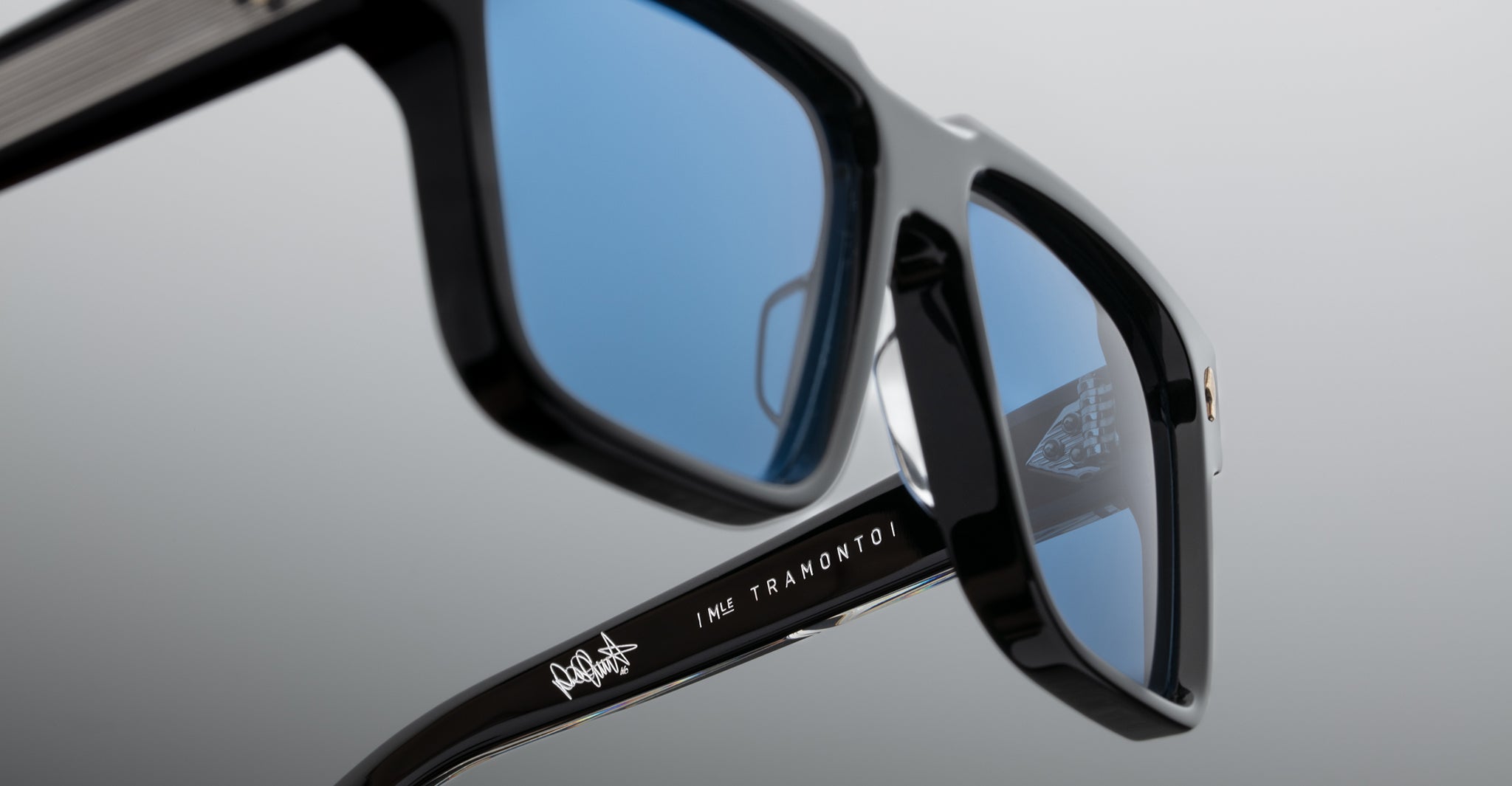 Close-up of black sunglasses with blue-tinted lenses. The arm of the sunglasses features white text and a small graphic design. The background is a smooth gradient from gray to white.