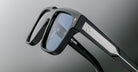 Close-up of a pair of sleek black sunglasses with blue-tinted lenses and silver detailing on the arms, shown against a blurred gray background.