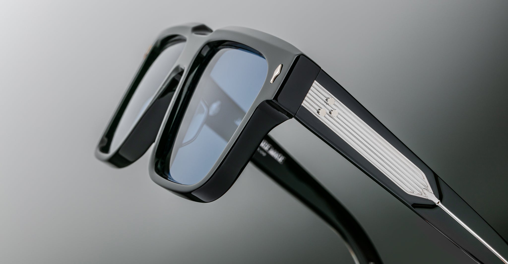 Close-up of a pair of sleek black sunglasses with blue-tinted lenses and silver detailing on the arms, shown against a blurred gray background.