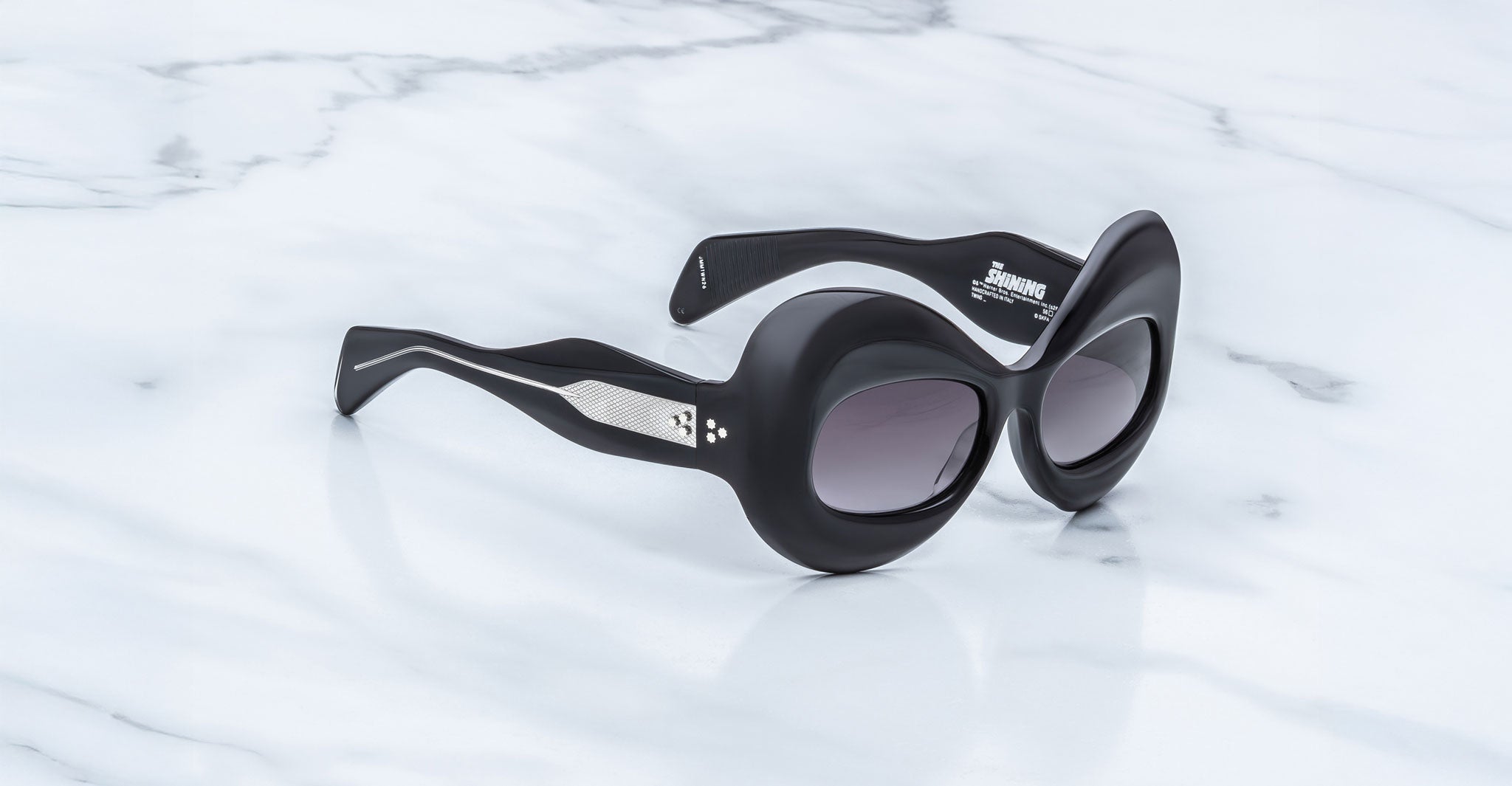 The Jacques Marie Mage Twins are black, oval sunglasses from the limited-edition The Shining Collection, inspired by Stanley Kubrick, with thick frames, wavy arms, and silver triangular accents displayed on a white marble surface.