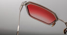 A close-up of the Jacques Marie Mage Ugo sunglasses with a metallic frame and red-tinted lenses, highlighting the hinge, nose pads, and side arm against a plain grey background.