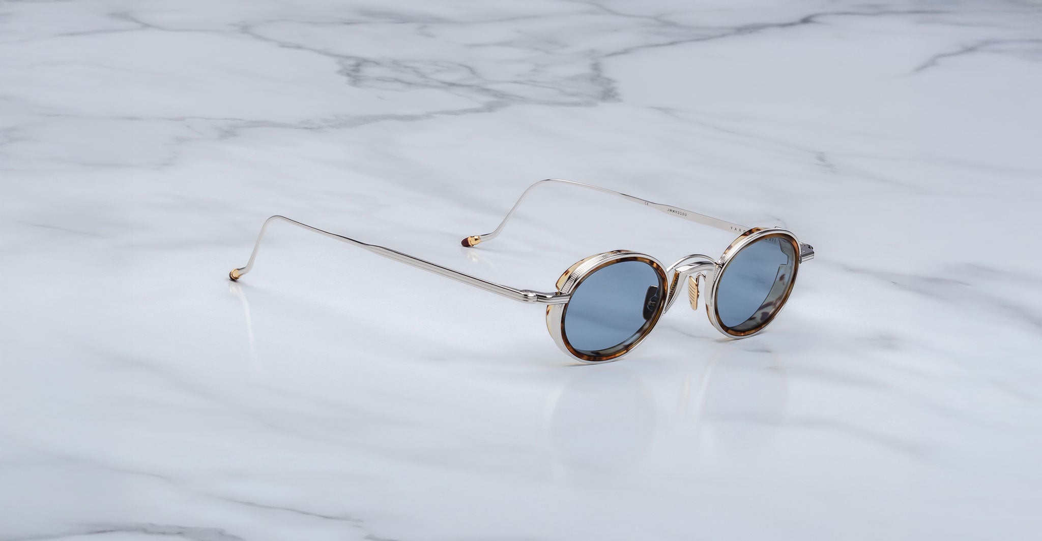 The Jacques Marie Mage Varda round sunglasses with thin gold frames and blue-tinted lenses rest on a white marble surface.
