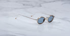 The Jacques Marie Mage Varda round sunglasses with thin gold frames and blue-tinted lenses rest on a white marble surface.