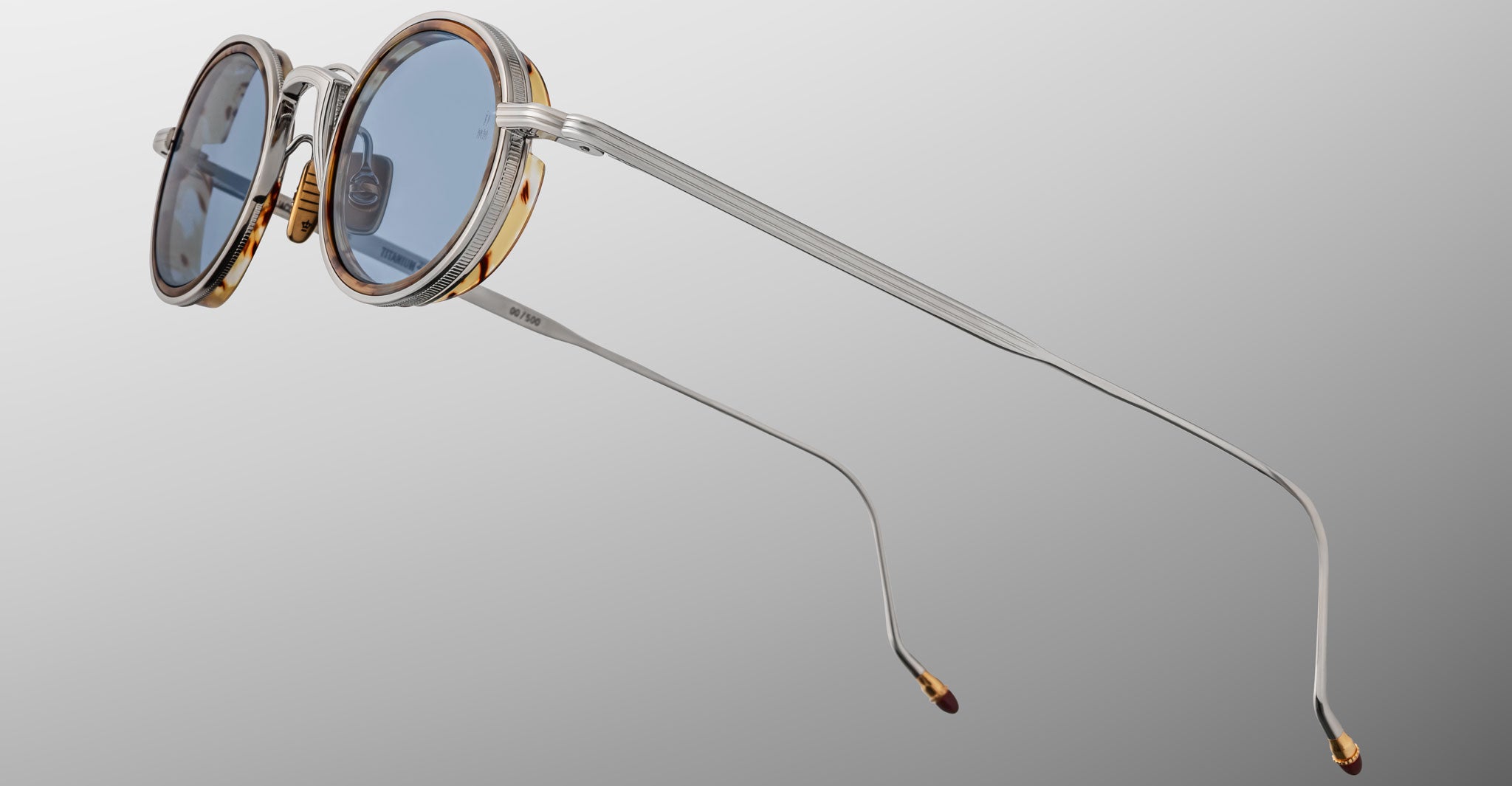 The Jacques Marie Mage Varda sunglasses feature round blue-tinted lenses, silver metal arms, and gold accents on the frame, displayed against a plain gradient background.
