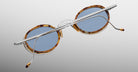 The Jacques Marie Mage Varda round sunglasses feature blue lenses, tortoiseshell frames, and thin silver metal arms, displayed from the back against a light gray gradient background.