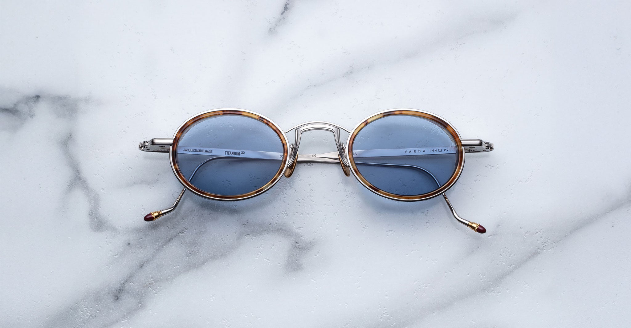 Jacques Marie Mage Varda round sunglasses with thin metal frames and blue-tinted lenses rest on white marble with light grey veining. The arms feature small decorative tips.