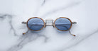 Jacques Marie Mage Varda round sunglasses with thin metal frames and blue-tinted lenses rest on white marble with light grey veining. The arms feature small decorative tips.