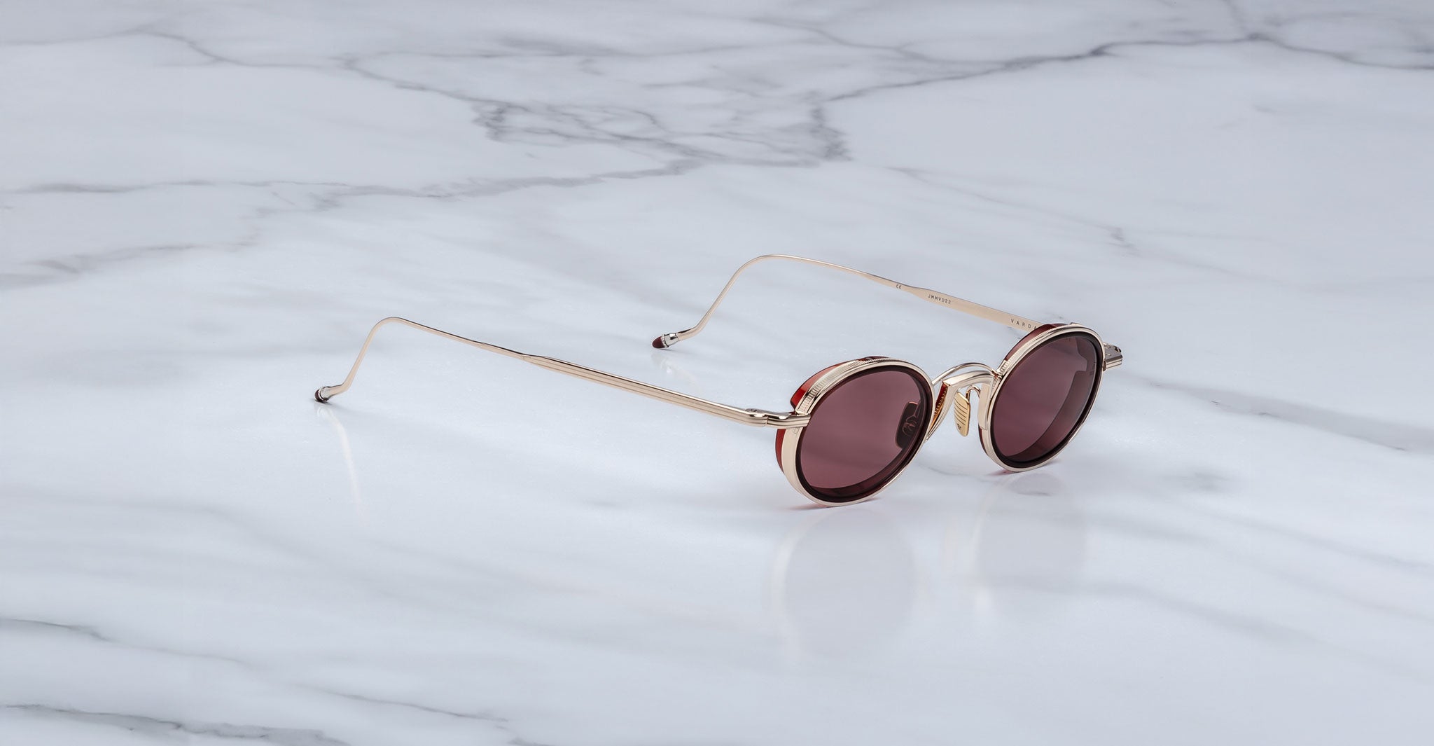 A pair of Jacques Marie Mage Varda round gold-framed sunglasses with dark tinted lenses rests on a white marble surface featuring gray veining.