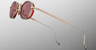 The Jacques Marie Mage Varda round sunglasses feature gold metal frames, thin gold arms, and pink-tinted lenses, displayed against a gray gradient background.