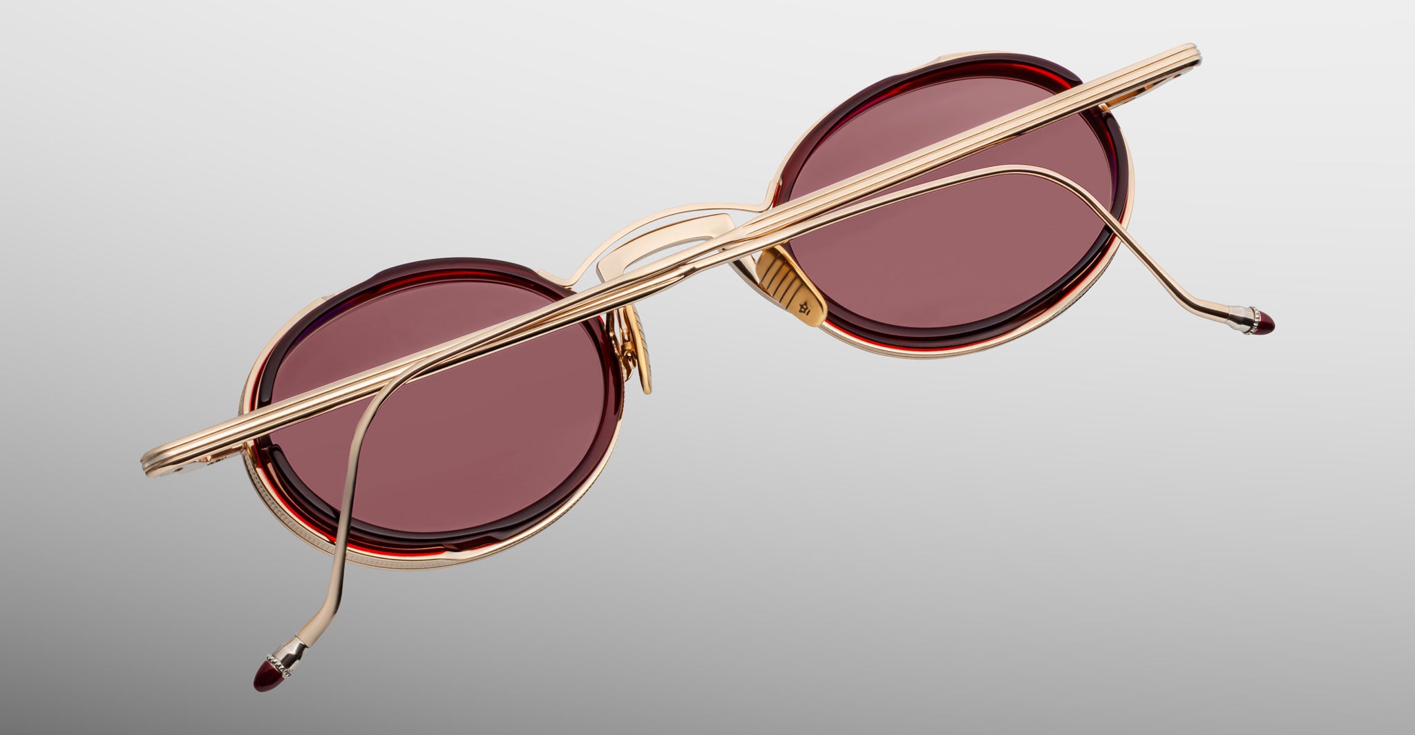 The Jacques Marie Mage Varda features round sunglasses with red-tinted lenses and gold metal frames, displayed from the back with crossed arms against a light grey background.