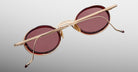 The Jacques Marie Mage Varda features round sunglasses with red-tinted lenses and gold metal frames, displayed from the back with crossed arms against a light grey background.