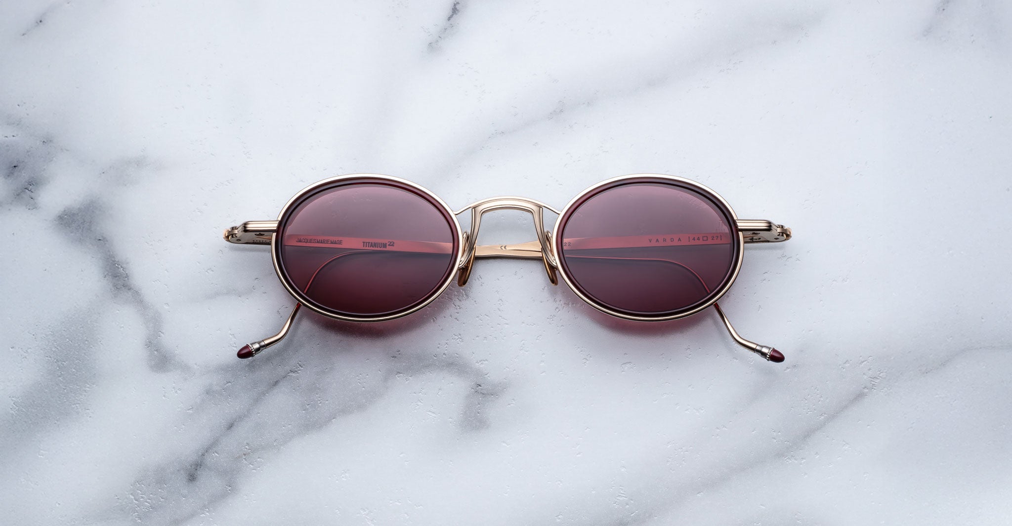 Jacques Marie Mage Varda round sunglasses with gold frames and dark pink tinted lenses rest on a white marble surface, their folded arms and the marbles reflection visible in the lenses.