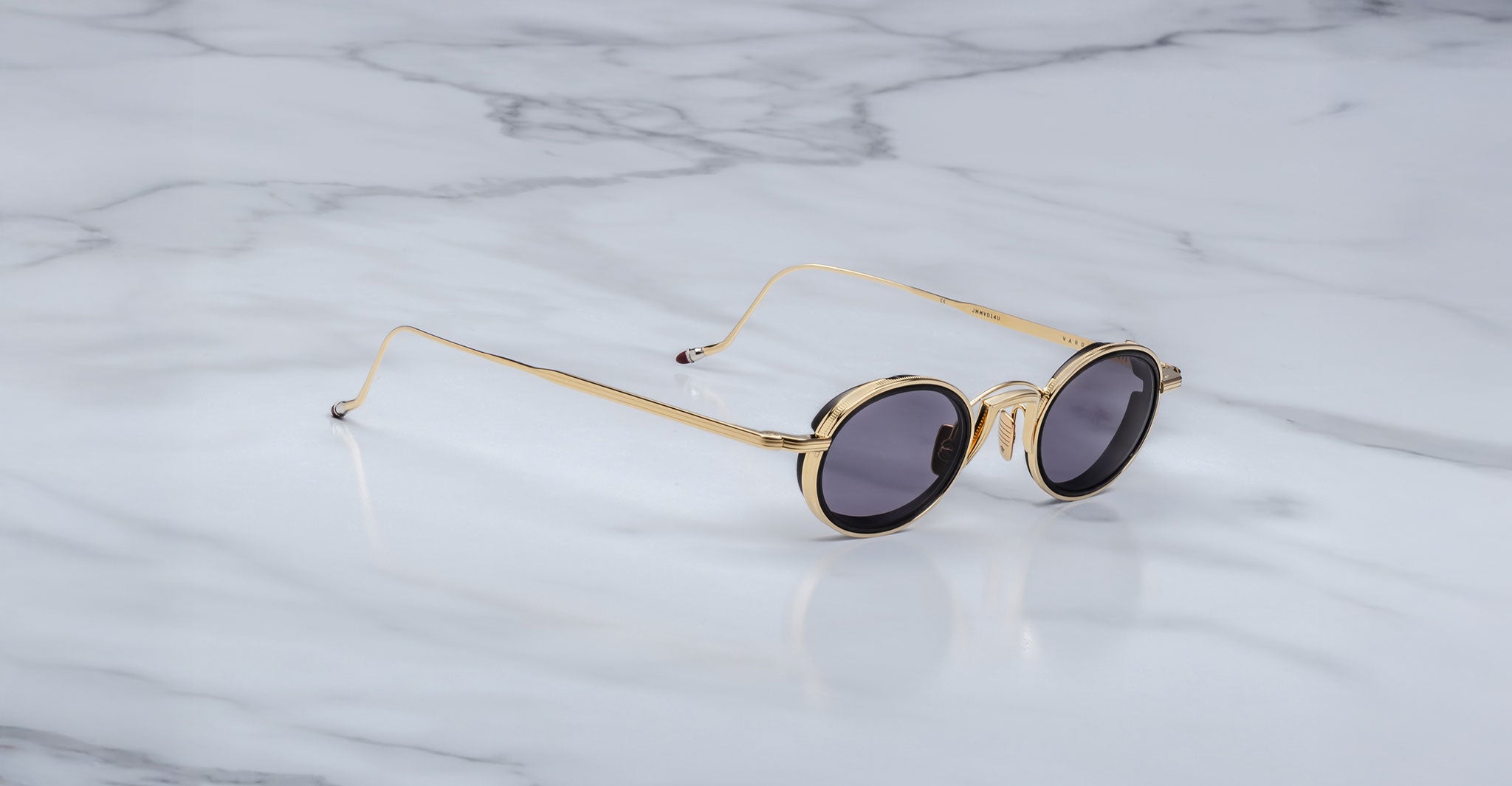 The Jacques Marie Mage Varda gold-framed round sunglasses with dark lenses are displayed on a white marble surface with gray veining.