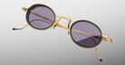 A pair of Jacques Marie Mage Varda round sunglasses with gold metal frames and dark tinted lenses shown against a light gray background. The temples feature a gold finish with distinctive small red tips.