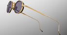 The Jacques Marie Mage Varda sunglasses feature round gold metal frames with black rims and dark tinted lenses, displayed at an angle on a neutral gray background. The thin temples are slightly curved at the tips.