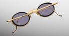 The Jacques Marie Mage Varda sunglasses, featuring round gold metal frames and dark lenses, are displayed from the back with folded temples resting on a light gray background.
