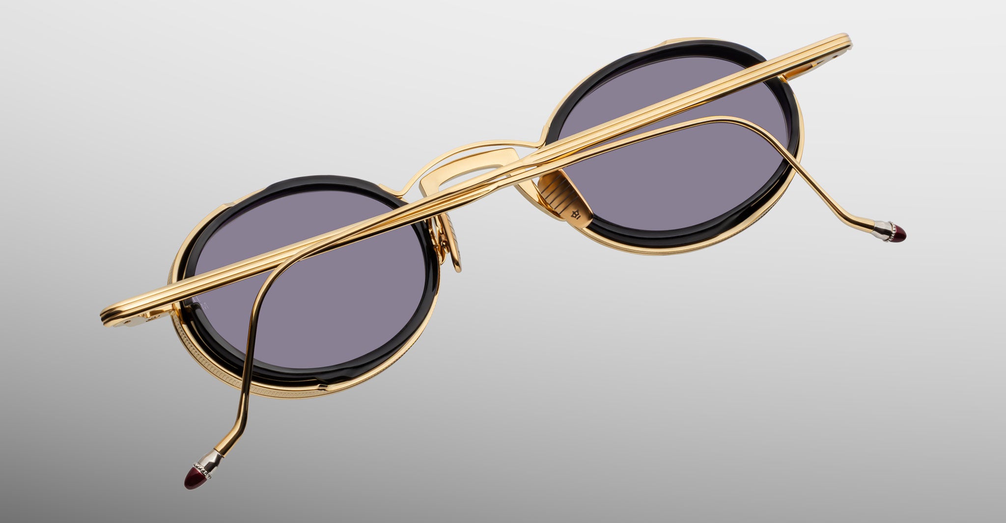 The Jacques Marie Mage Varda sunglasses, featuring round gold metal frames and dark lenses, are displayed from the back with folded temples resting on a light gray background.