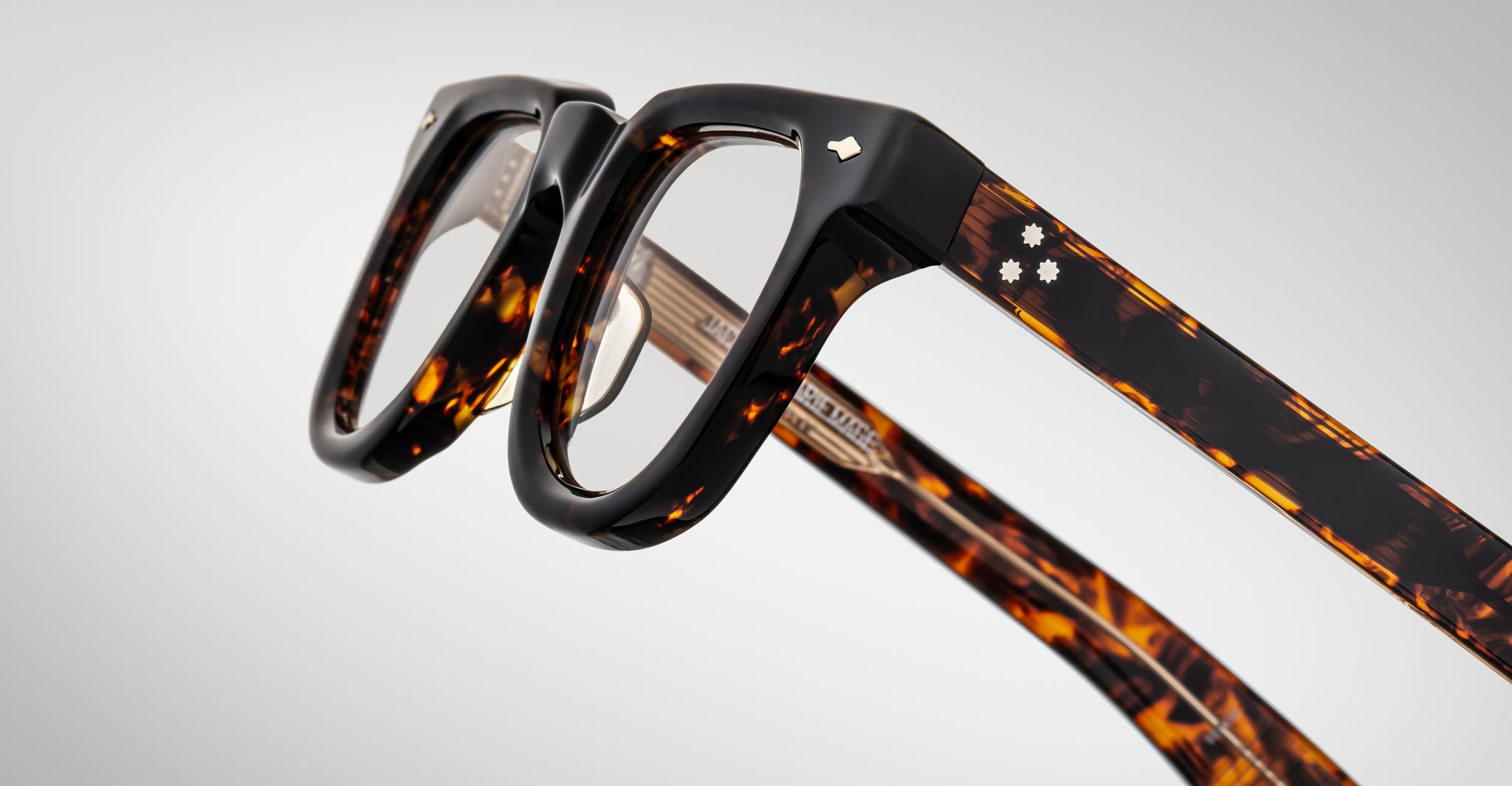 Close-up of Jacques Marie Mage Vendome optical frames in tortoiseshell with thick arms and metallic accents, handcrafted in Japan, displayed on a plain gray background.