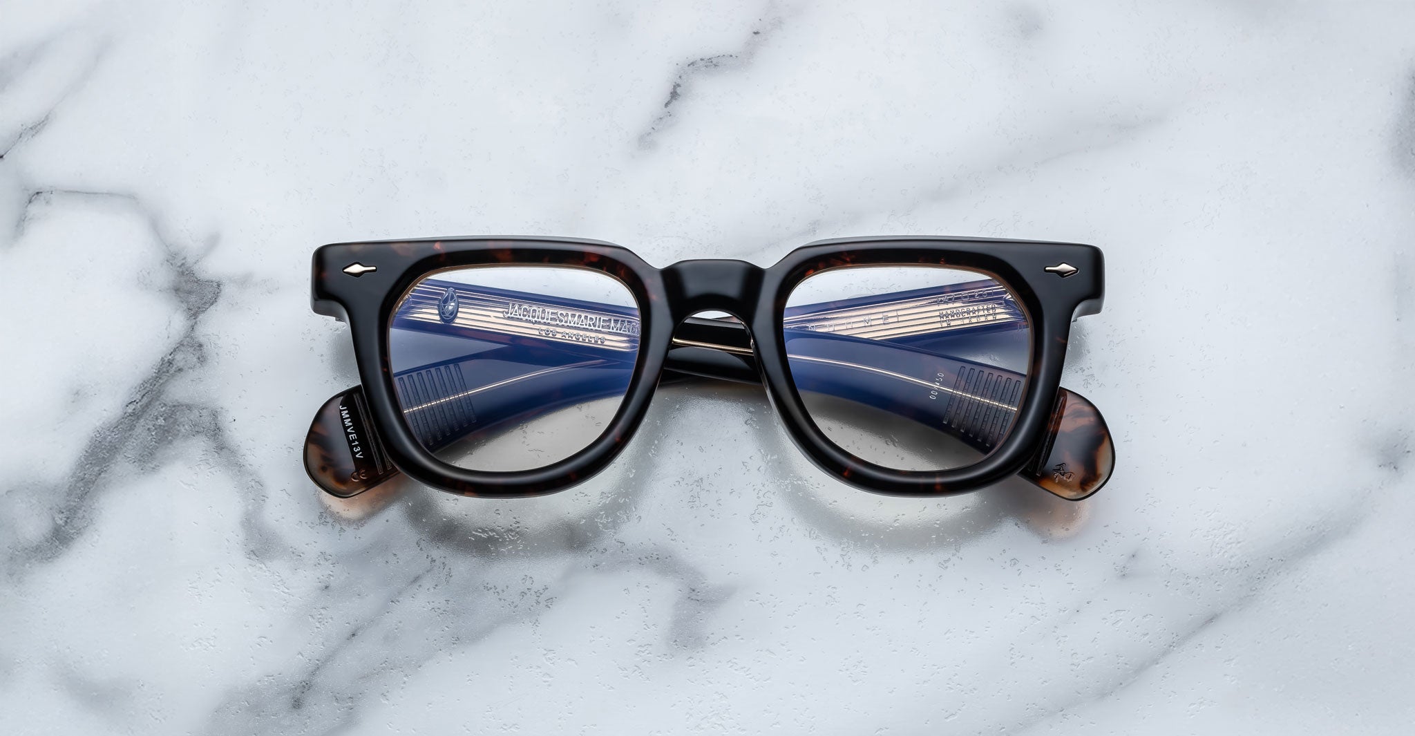 A pair of Jacques Marie Mage Vendome frames by Jacques Marie Mage sits on a white marble surface, their lenses reflecting a blue and white building.