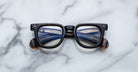A pair of Jacques Marie Mage Vendome frames by Jacques Marie Mage sits on a white marble surface, their lenses reflecting a blue and white building.