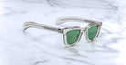 Jacques Marie Mage Vendome sunglasses by Jacques Marie Mage, featuring clear frames and green lenses, handcrafted in Japan, rest on a white marble surface.