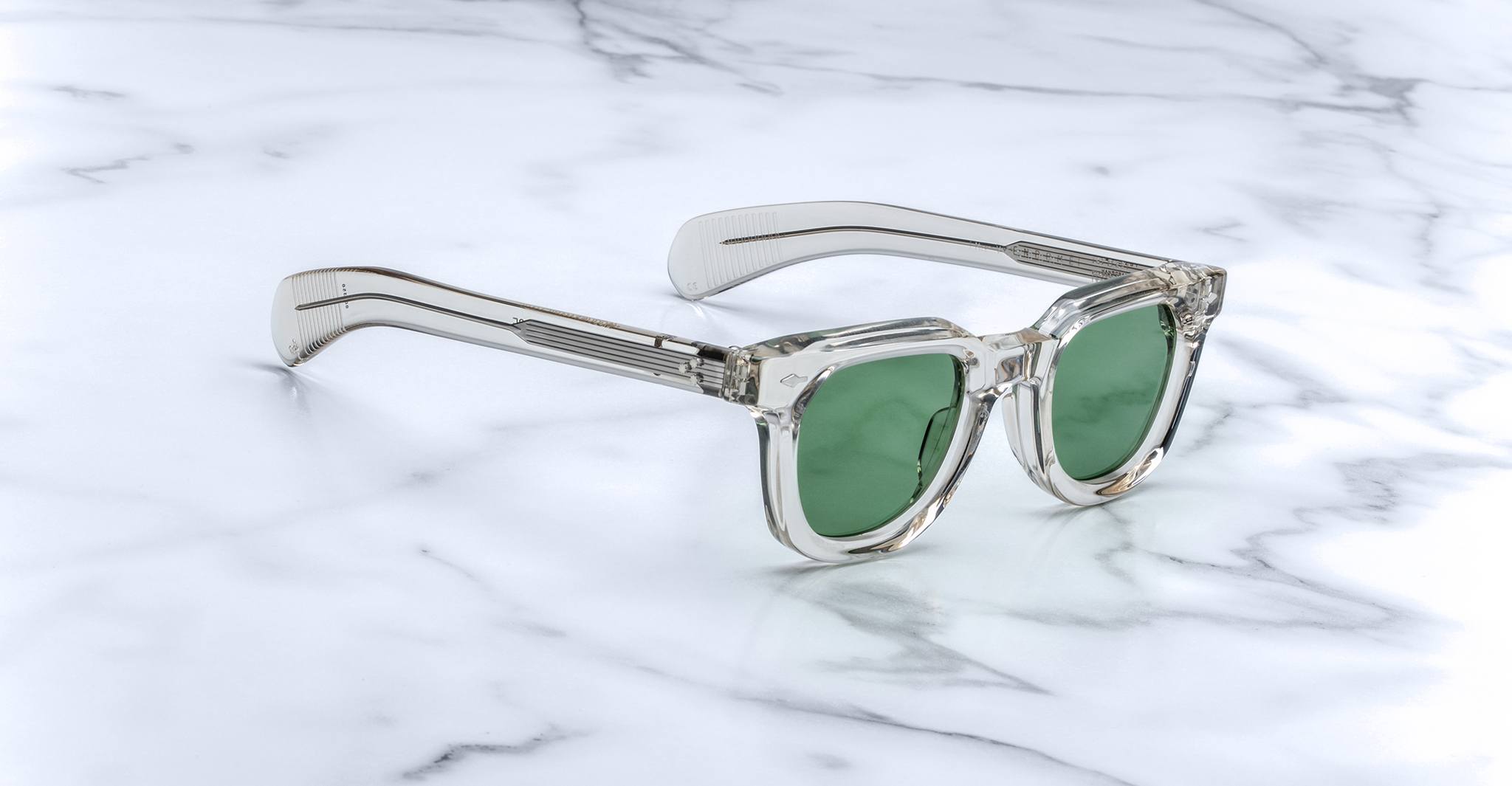 Jacques Marie Mage Vendome sunglasses by Jacques Marie Mage, featuring clear frames and green lenses, handcrafted in Japan, rest on a white marble surface.