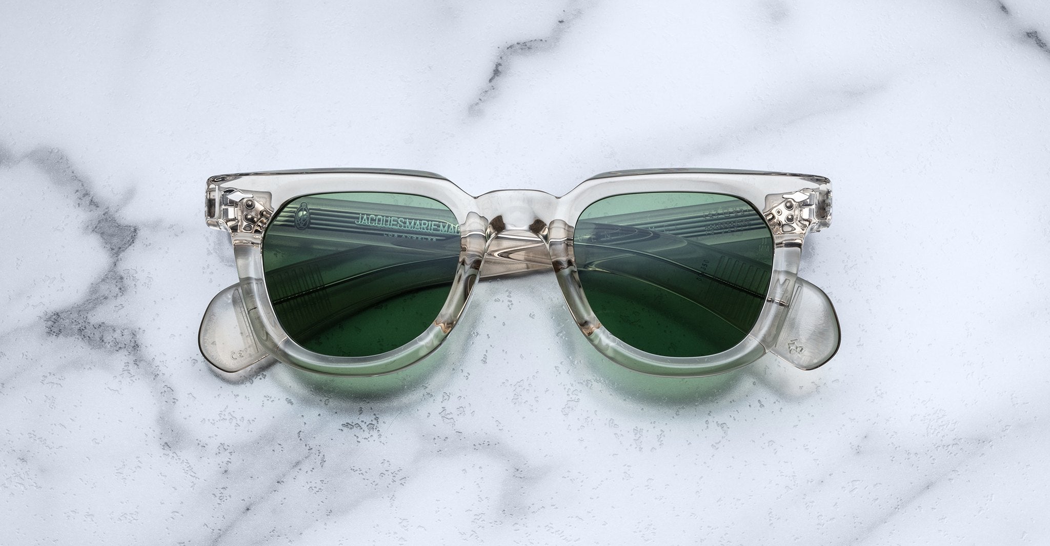 Jacques Marie Mage Vendome clear-frame sunglasses with green lenses, handcrafted in Japan by Jacques Marie Mage, displayed on a white marble surface.