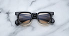 A pair of Jacques Marie Mage Vendome sunglasses by Jacques Marie Mage, featuring black rims and brown gradient lenses, handcrafted in Japan, is displayed on a white marble surface.