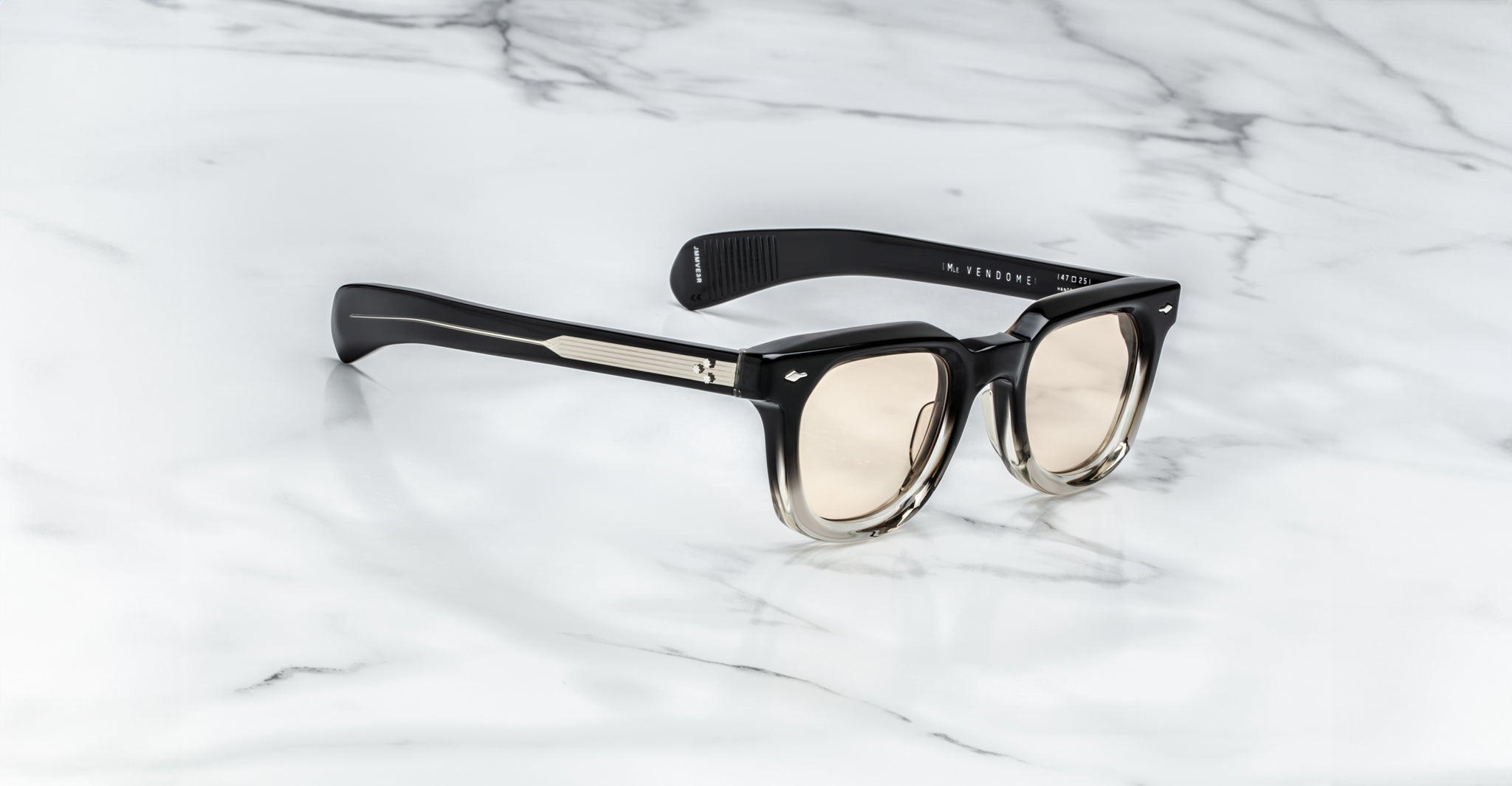 Jacques Marie Mage Vendome sunglasses in black and clear with light-tinted lenses sit on white marble, featuring handcrafted Japanese optical frames by Jacques Marie Mage.
