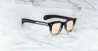 Jacques Marie Mage Vendome sunglasses in black and clear with light-tinted lenses sit on white marble, featuring handcrafted Japanese optical frames by Jacques Marie Mage.