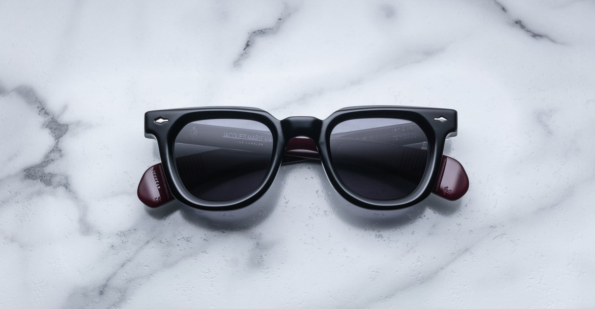Jacques Marie Mage Vendome sunglasses featuring black square frames, dark lenses, and burgundy arms rest on a white marble surface. Handcrafted in Japan by Jacques Marie Mage.