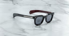 The Jacques Marie Mage Vendome sunglasses, by Jacques Marie Mage, are black rectangular frames with dark lenses, silver arm detailing, and maroon inner temples—handcrafted in Japan and displayed on a white marble surface.
