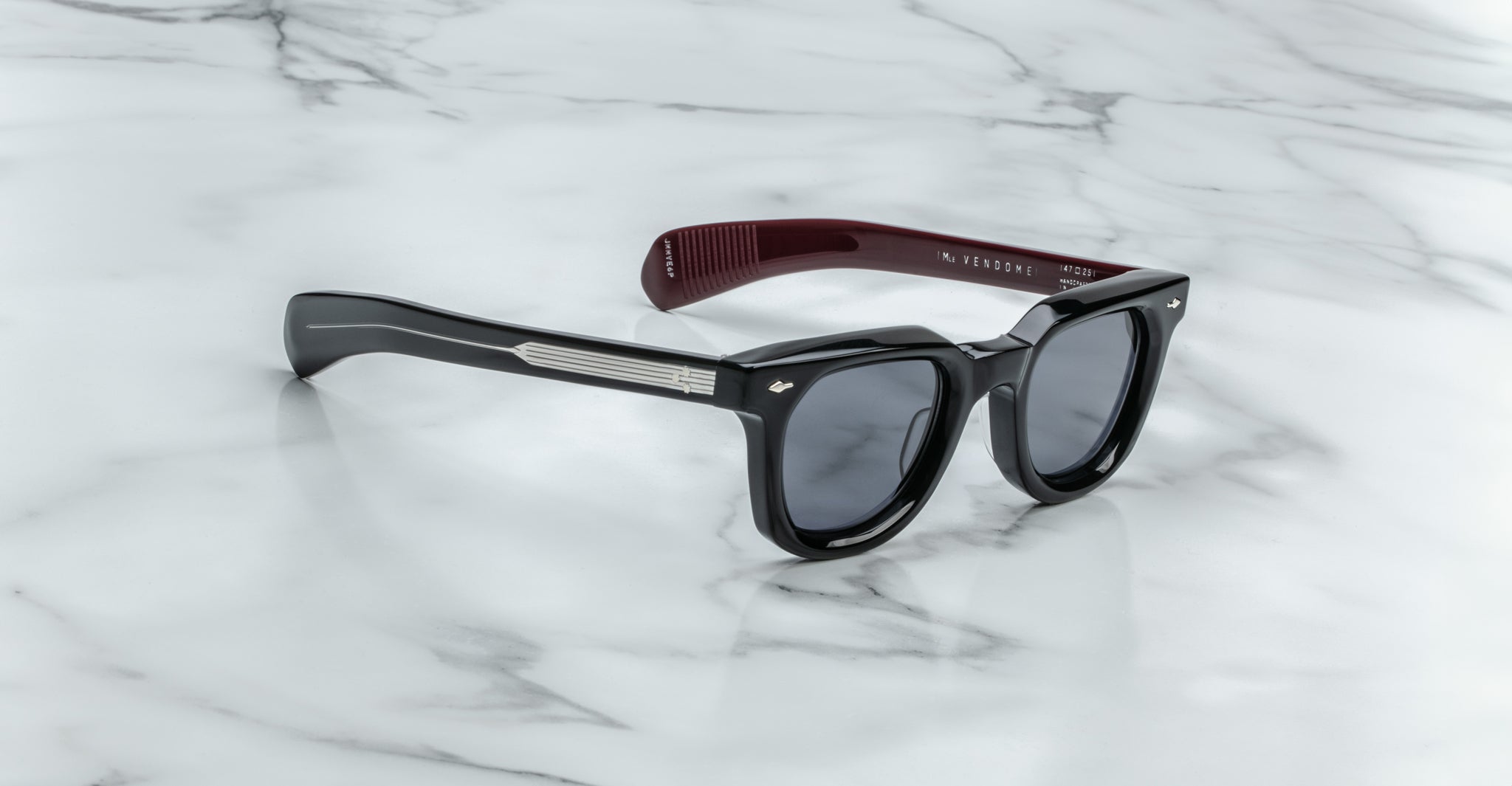 The Jacques Marie Mage Vendome sunglasses, by Jacques Marie Mage, are black rectangular frames with dark lenses, silver arm detailing, and maroon inner temples—handcrafted in Japan and displayed on a white marble surface.