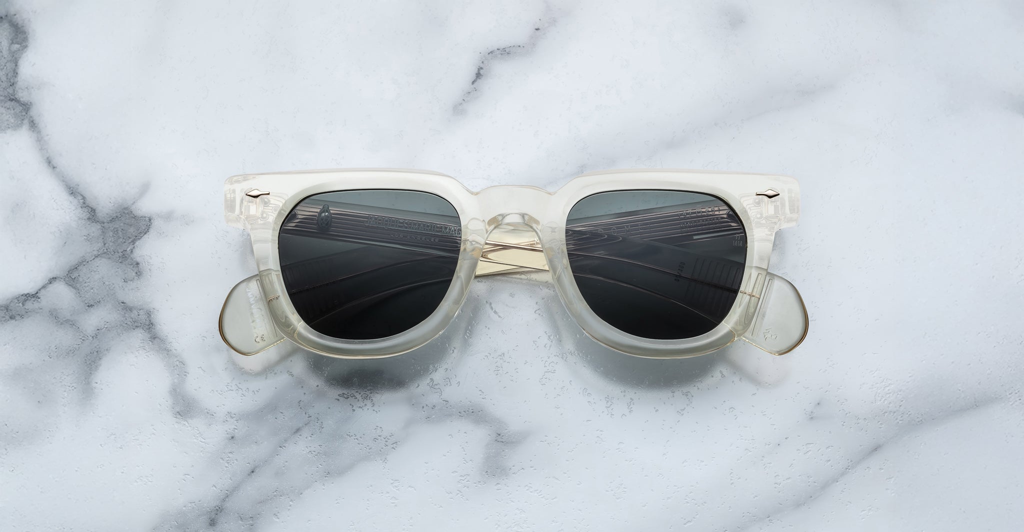 Jacques Marie Mage Vendome sunglasses by Jacques Marie Mage, featuring clear frames and dark lenses, handcrafted in Japan, displayed on a white and gray marble surface.