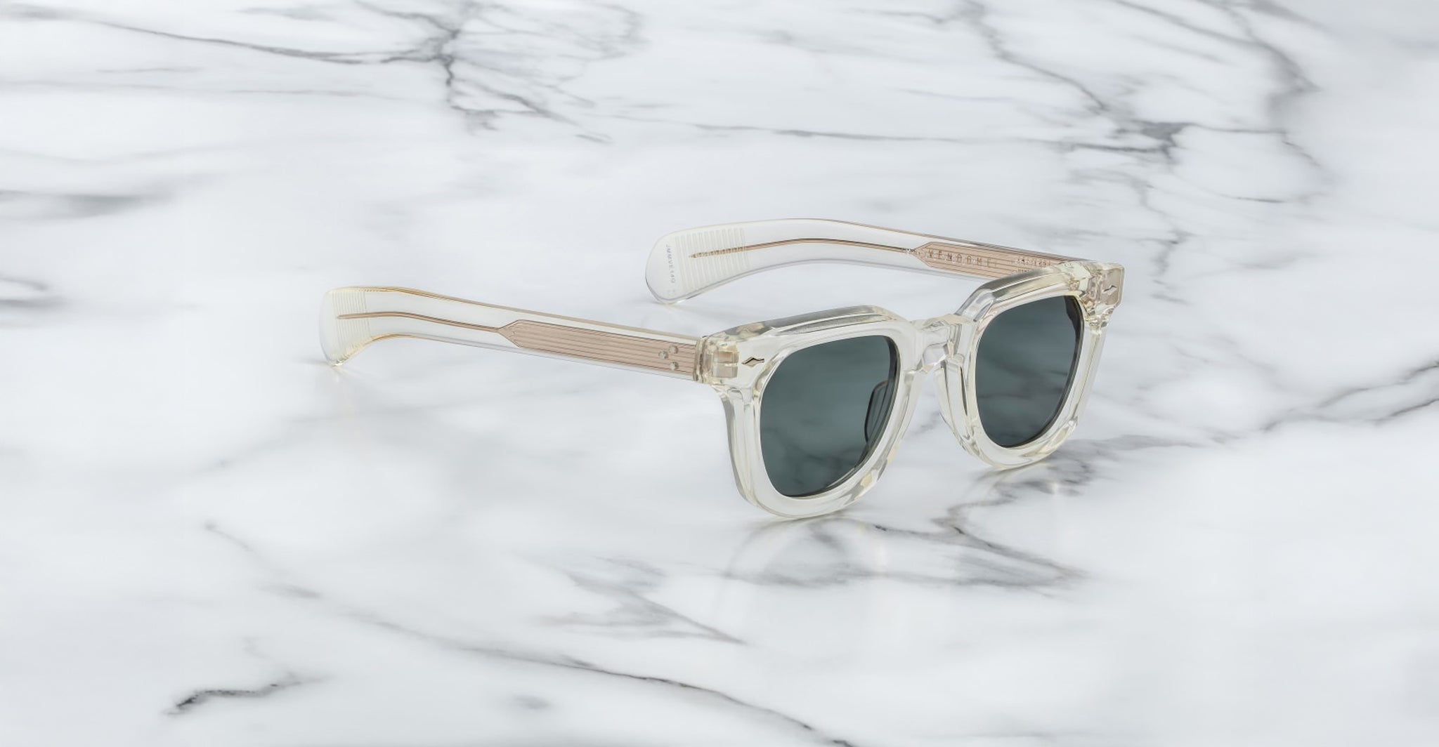Jacques Marie Mage Vendome sunglasses with clear frames, dark lenses, and gold-tone temples by Jacques Marie Mage are displayed on a white marble surface.