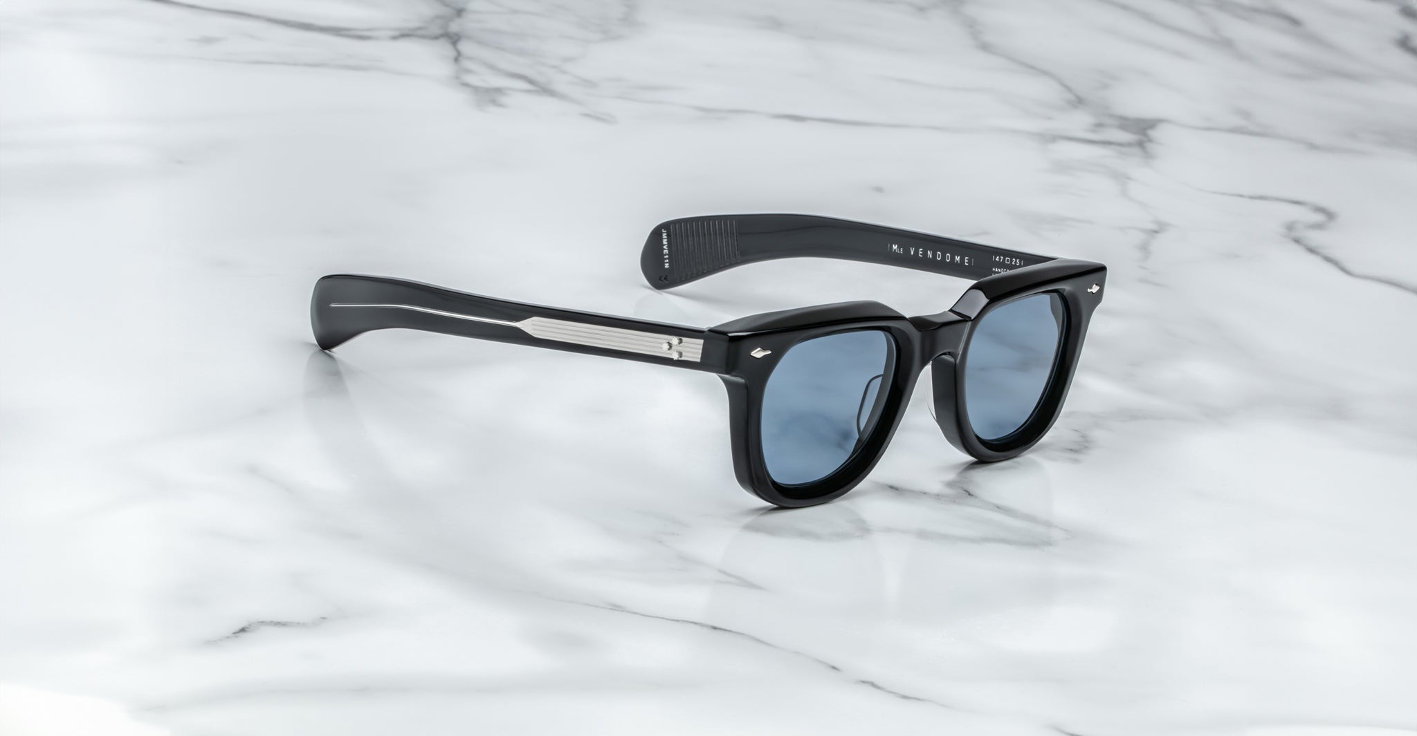 A pair of Jacques Marie Mage Vendome sunglasses by Jacques Marie Mage, handcrafted in Japan with black rectangular frames and blue lenses, rests on a white marble surface.