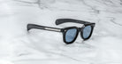 A pair of Jacques Marie Mage Vendome sunglasses by Jacques Marie Mage, handcrafted in Japan with black rectangular frames and blue lenses, rests on a white marble surface.
