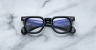 Jacques Marie Mage Vendome optical frames by Jacques Marie Mage, handcrafted in Japan, showcase black rectangular eyeglasses with blue-tinted lenses on a white marble surface and subtle lens reflections.
