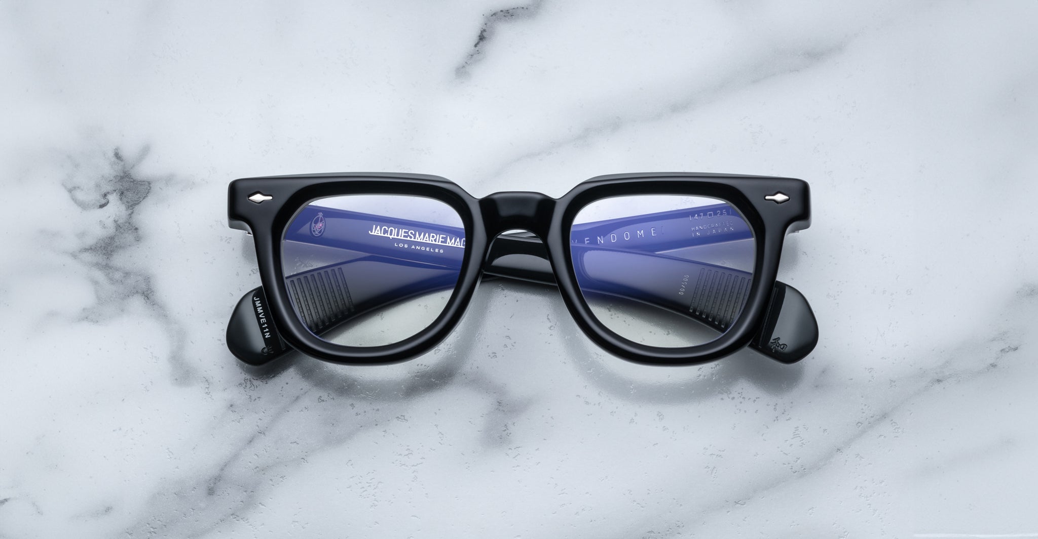 Jacques Marie Mage Vendome optical frames by Jacques Marie Mage, handcrafted in Japan, showcase black rectangular eyeglasses with blue-tinted lenses on a white marble surface and subtle lens reflections.