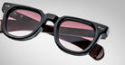 Close-up of Jacques Marie Mage Vendome sunglasses, featuring black frames and pink-tinted lenses, handcrafted in Japan. Shown at an angle on a neutral background with "VENDOME" visible on the inside of the arm.