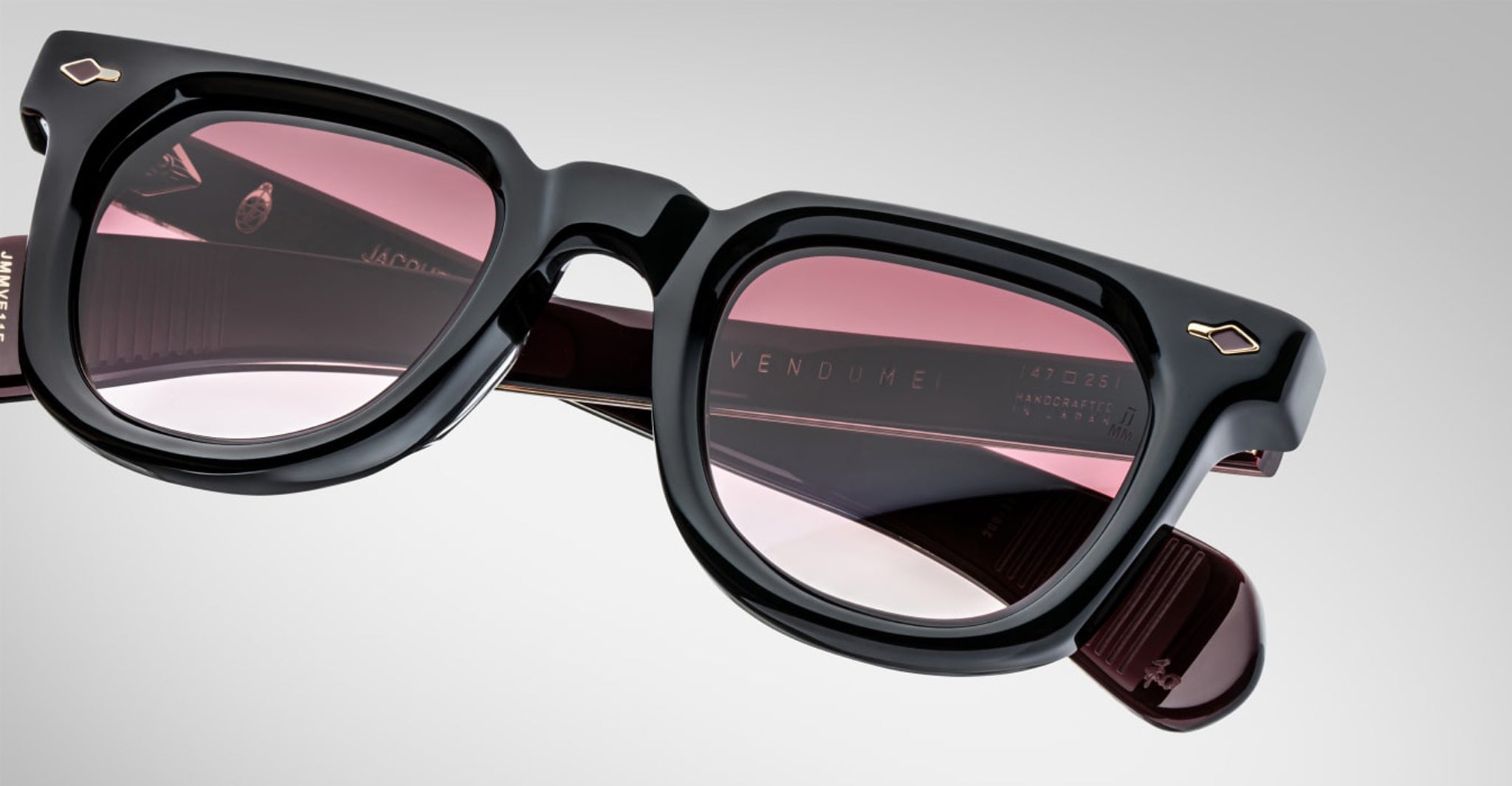Close-up of Jacques Marie Mage Vendome sunglasses, featuring black frames and pink-tinted lenses, handcrafted in Japan. Shown at an angle on a neutral background with "VENDOME" visible on the inside of the arm.