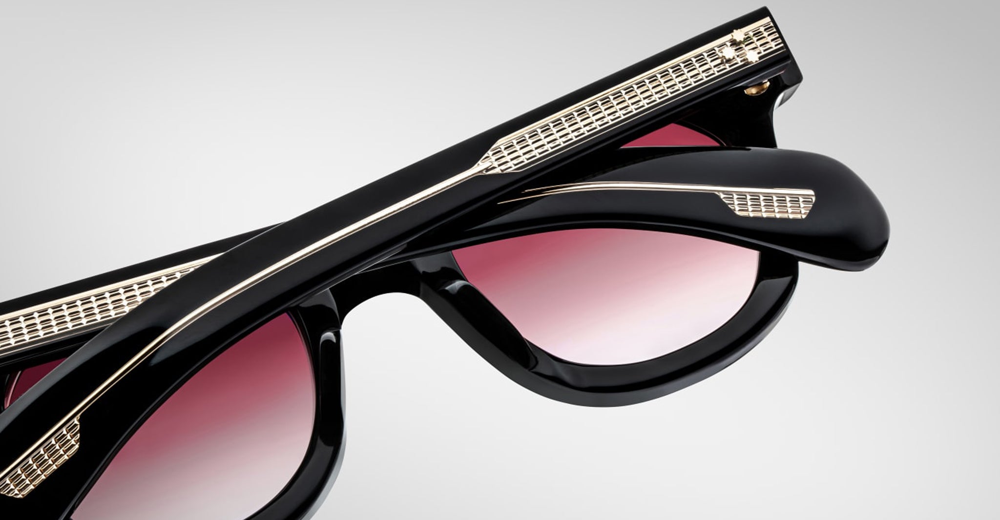 A close-up of Jacques Marie Mage Vendome sunglasses by Jacques Marie Mage, featuring black frames, gold detailing, and pink gradient lenses, handcrafted in Japan and shown folded on a plain light gray background.
