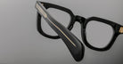 Close-up of Jacques Marie Mage Vendome optical frames in thick black Japanese acetate with gold temple detailing by Jacques Marie Mage, set against a plain gray background.