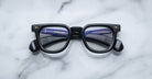 A pair of Jacques Marie Mage Vendome black optical frames, featuring a thick handcrafted-in-Japan design, rests on white marble with the Jacques Marie Mage brand information reflected in the lenses.