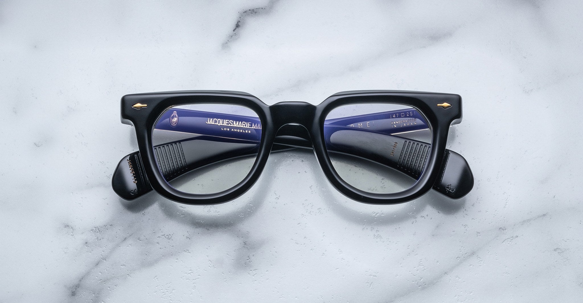 A pair of Jacques Marie Mage Vendome black optical frames, featuring a thick handcrafted-in-Japan design, rests on white marble with the Jacques Marie Mage brand information reflected in the lenses.