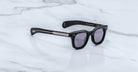 The Jacques Marie Mage Vendome sunglasses by Jacques Marie Mage feature black rectangular thick frames with silver temple detailing, handcrafted in Japan, displayed on a white marble surface.