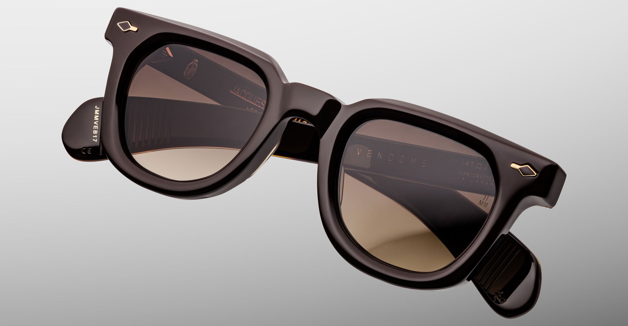 A pair of Jacques Marie Mage Vendome sunglasses by Jacques Marie Mage, featuring thick glossy black frames and brown tinted lenses, handcrafted in Japan and displayed at an angle on a light grey background.